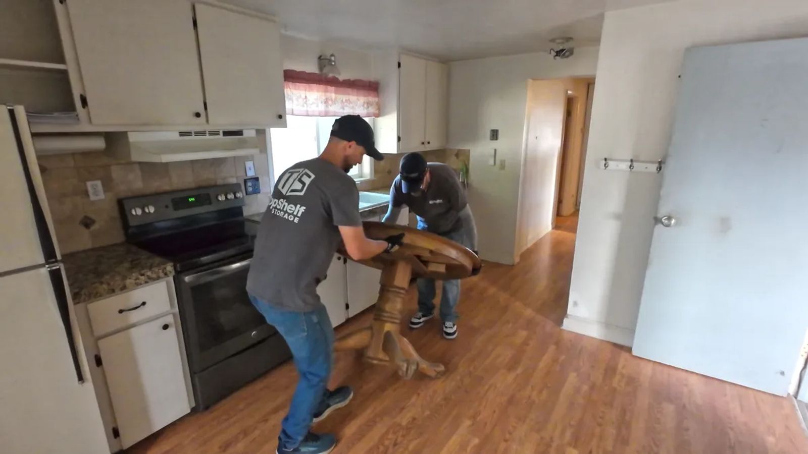 Two Top Shelf crew members in branded shirts tipping a heavy wooden pedestal table on its side during a kitchen furniture removal in Eagle Idaho