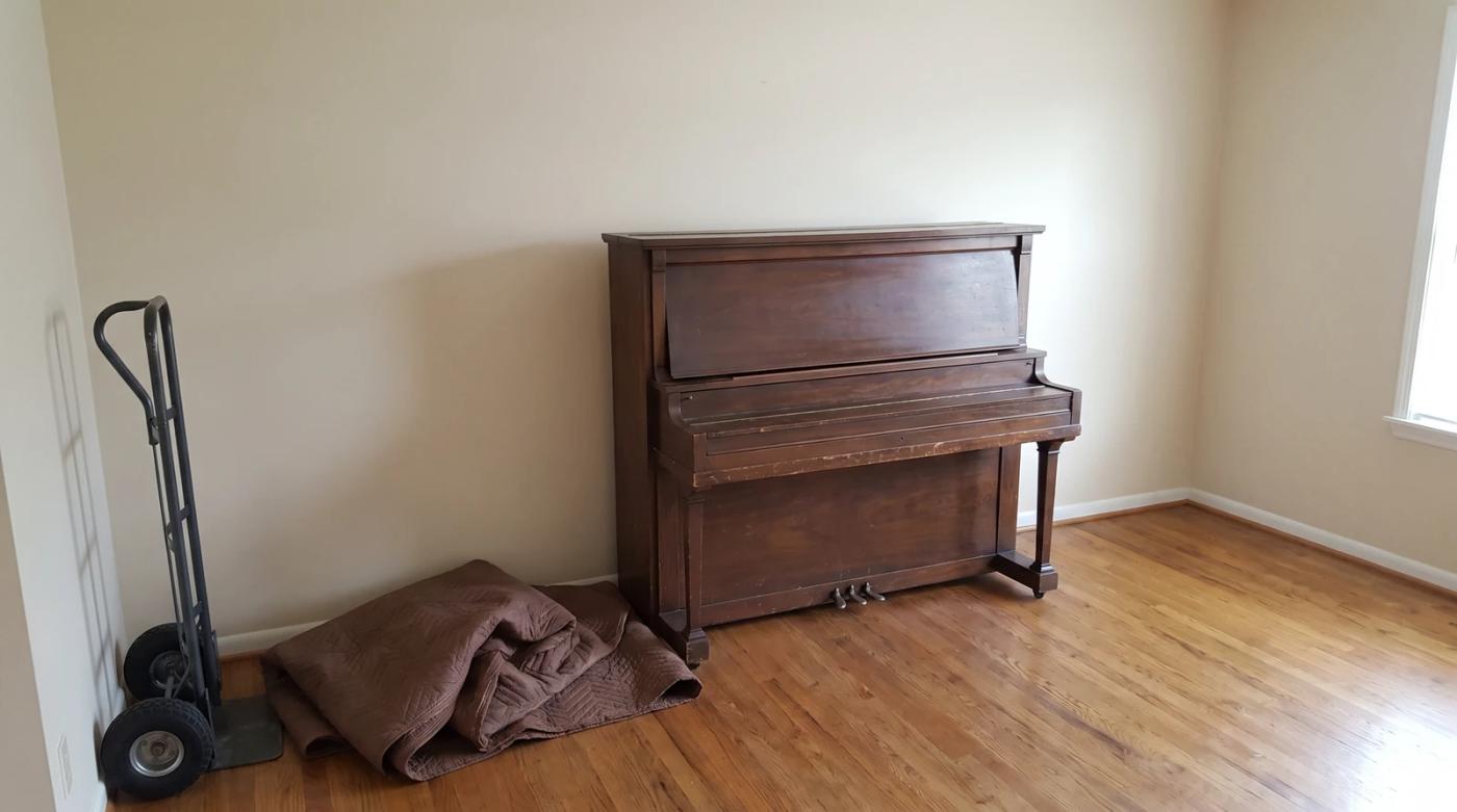 Old upright piano waiting for removal in a Garden City, Idaho living room, with a folded moving blanket and a furniture dolly beside it