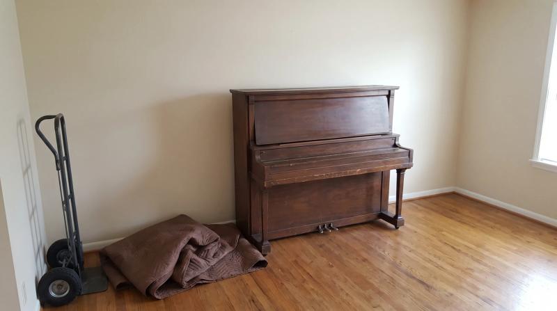 Old upright piano waiting for removal in a Garden City, Idaho living room, with a folded moving blanket and a furniture dolly beside it