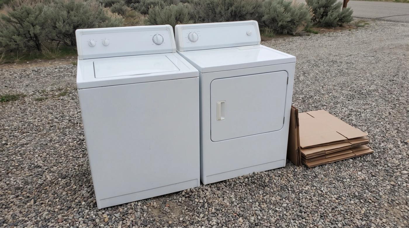 An old washer and dryer set on a gravel driveway beside stacked cardboard, waiting for appliance removal at a home in Star, Idaho.