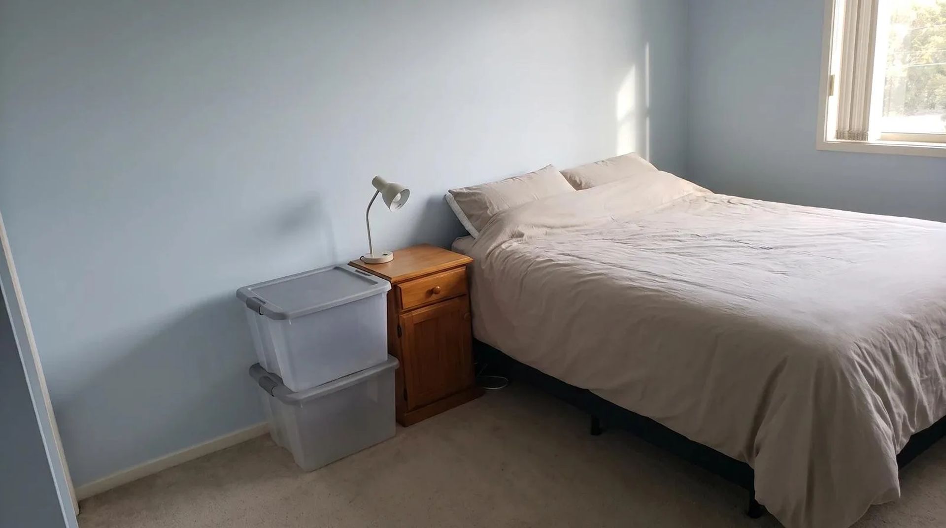 A neatly made bed in a bare bedroom with a small lamp on the nightstand and two empty plastic bins stacked beside it, in a Boise home during a residential hoarding cleanup