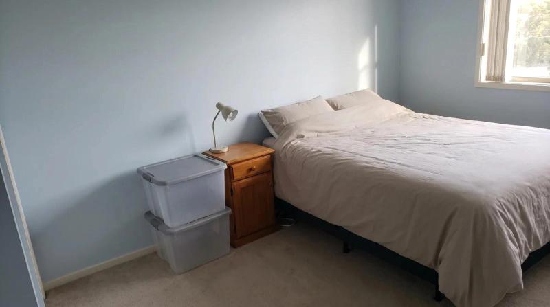 A neatly made bed in a bare bedroom with a small lamp on the nightstand and two empty plastic bins stacked beside it, in a Boise home during a residential hoarding cleanup