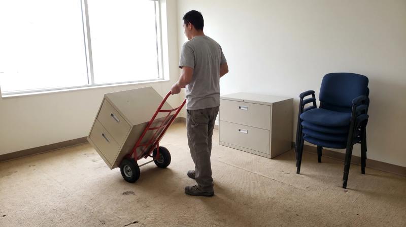 Worker moving a metal filing cabinet on a hand dolly during an office cleanout in a mostly empty office suite in Caldwell, Idaho