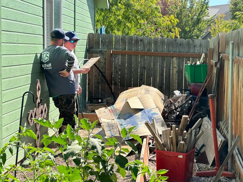 Two Top Shelf crew members assessing a pile of cardboard broken fence pieces and yard debris along the side of a house during a yard cleanout in Emmett Idaho