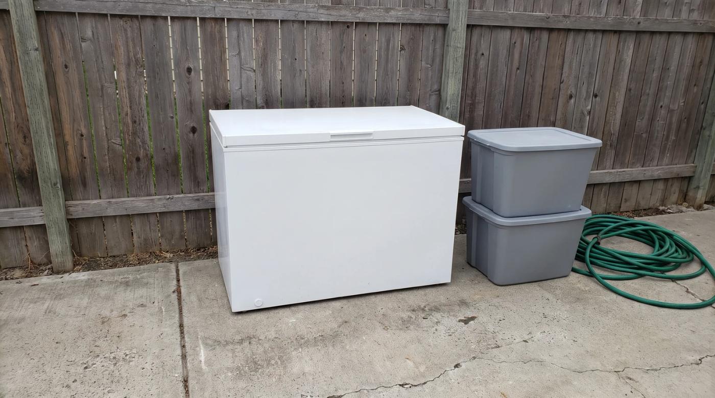 Old white chest freezer set out on a concrete driveway next to stacked storage totes at a home in Eagle, Idaho, ready for appliance removal.