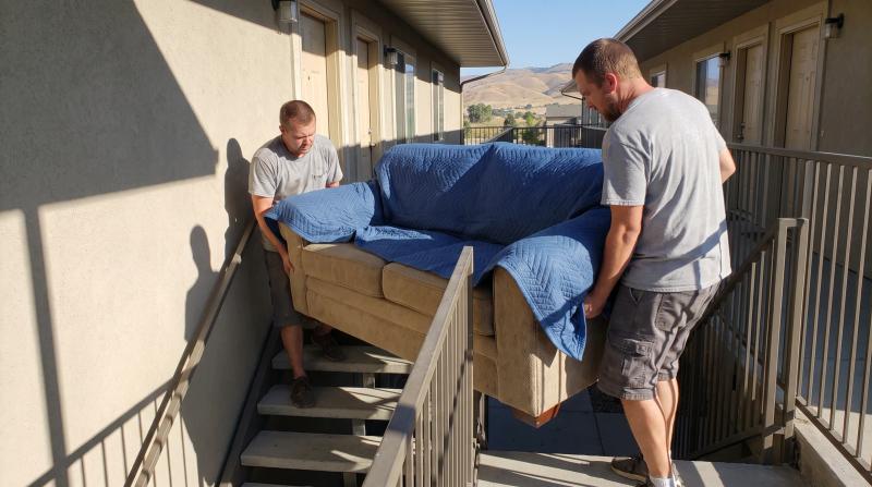 Two movers carrying an old sofa wrapped in a moving blanket down apartment stairs during a couch removal in Nampa, Idaho