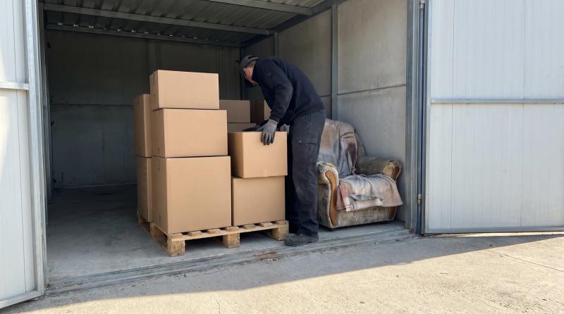 A person in plain work clothes lifting a plain cardboard box from an open storage unit stacked with boxes and an old armchair near Eagle, Idaho.