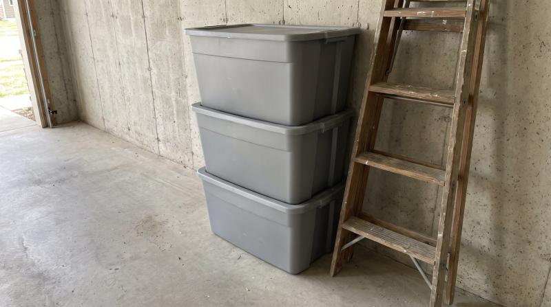 Stacked plastic storage totes and a leaning wooden ladder against a bare concrete wall in a Boise area garage before a cleanout