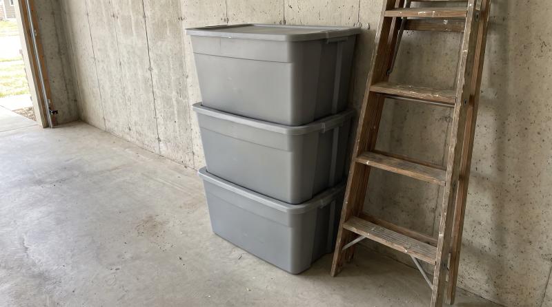 Stacked plastic storage totes and a leaning wooden ladder against a bare concrete wall in a Boise area garage before a cleanout