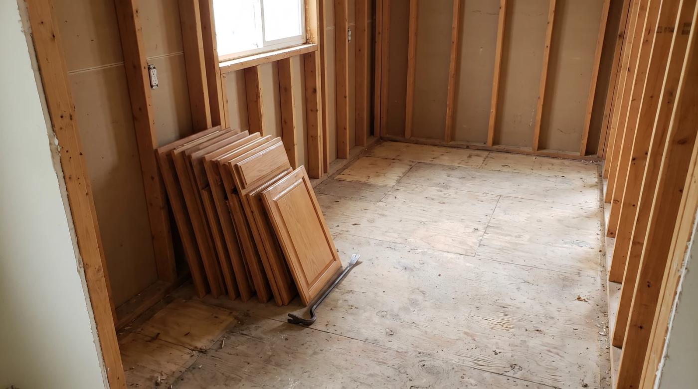 Kitchen stripped to bare studs and plywood subfloor during an interior demolition in Boise, Idaho, with removed cabinet doors stacked against the wall and a pry bar on the floor