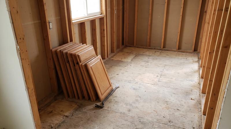 Kitchen stripped to bare studs and plywood subfloor during an interior demolition in Boise, Idaho, with removed cabinet doors stacked against the wall and a pry bar on the floor