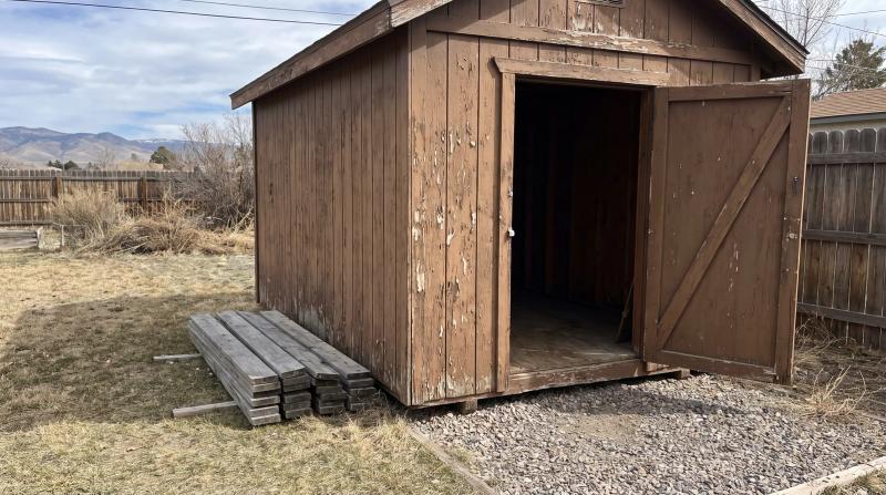 An empty weathered wooden shed with its door open and loose boards stacked beside it in a backyard in Caldwell, Idaho, cleared out and ready for demolition.