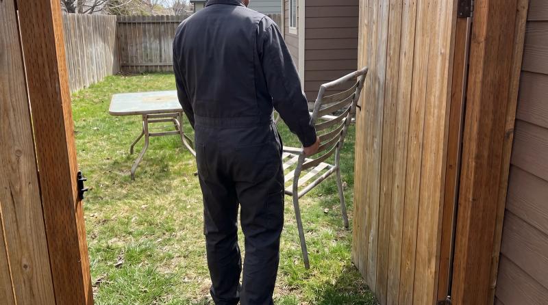 Worker carrying an old aluminum patio chair through a backyard side gate during patio furniture removal in Kuna, Idaho