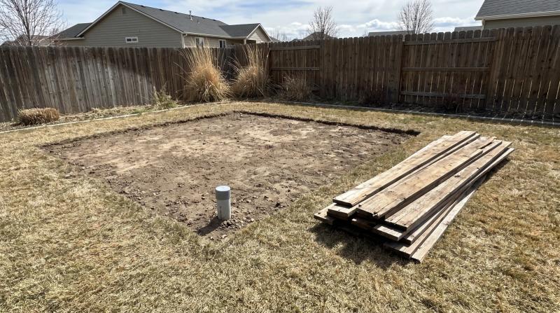 Bare dirt footprint and capped conduit stub left after a backyard shed demolition in Kuna, Idaho, with stacked wood siding boards on the grass