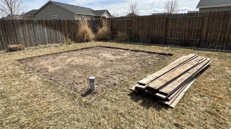 Bare dirt footprint and capped conduit stub left after a backyard shed demolition in Kuna, Idaho, with stacked wood siding boards on the grass