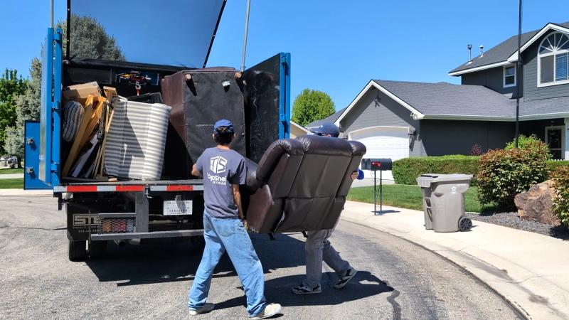 Two Top Shelf crew members in branded shirts lifting a brown leather recliner into the junk removal truck in a Meridian Idaho subdivision
