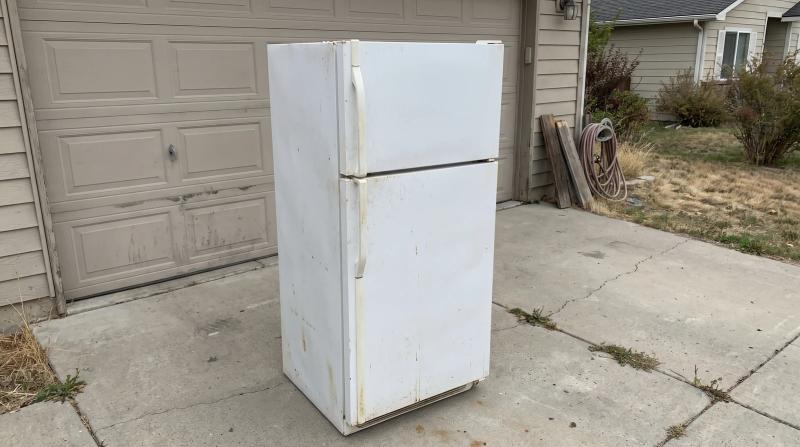 Two workers in plain clothes preparing to lift an old white refrigerator off a garage floor in Meridian, Idaho.