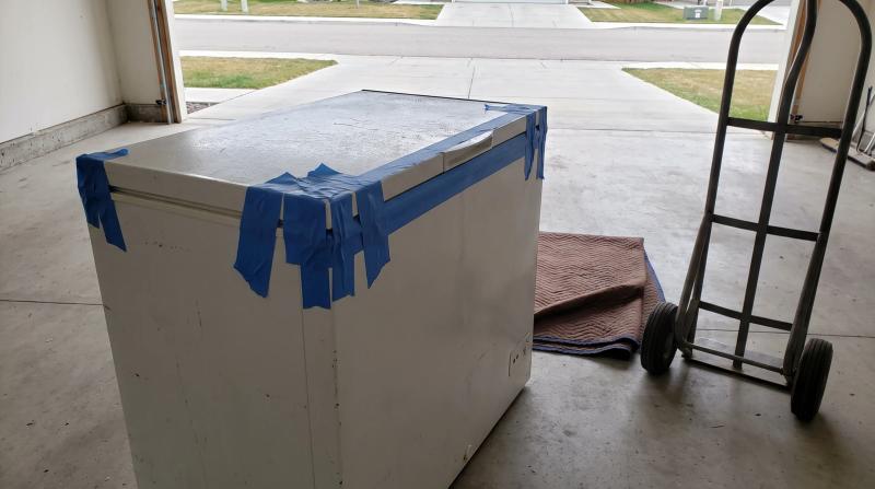 An old white chest freezer taped shut on a concrete garage floor in Star, Idaho, with a moving blanket and hand dolly waiting beside it for appliance removal.