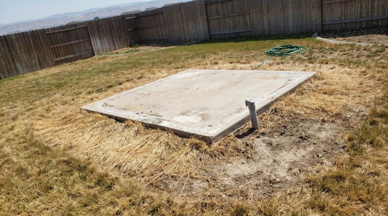 An empty concrete hot tub pad in a Caldwell, Idaho backyard, ringed by flattened yellow grass with a stub of electrical conduit at one corner