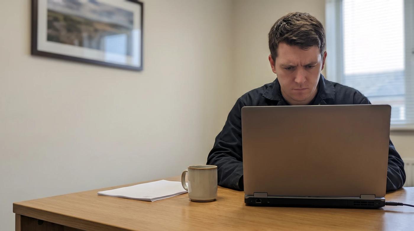 Small business owner at a desk looking at a laptop next to a coffee mug and a stack of business documents