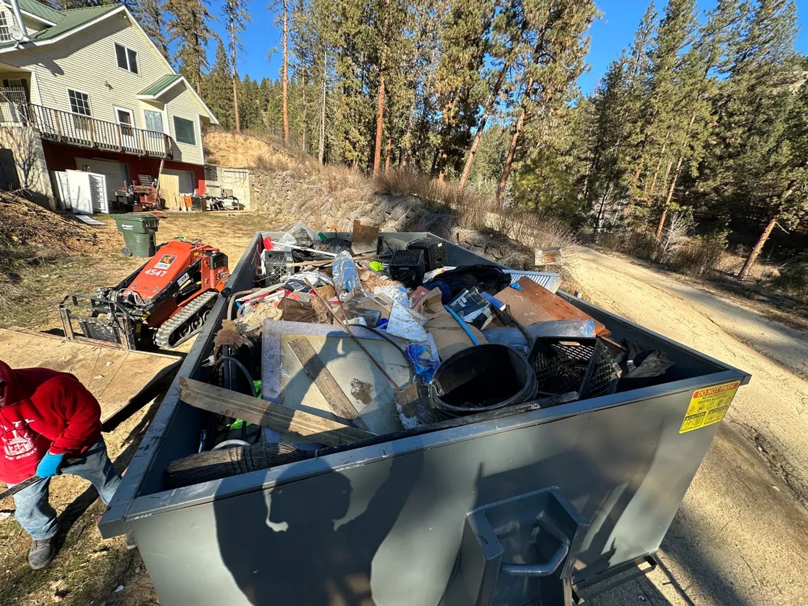 Top Shelf crew loading a full dumpster with property debris using a mini track loader during a hillside cleanout job in Star Idaho
