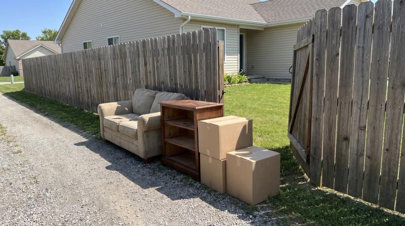 Old couch, wooden dresser and cardboard boxes stacked beside an open wooden gate at the end of a narrow gravel driveway in Boise, Idaho