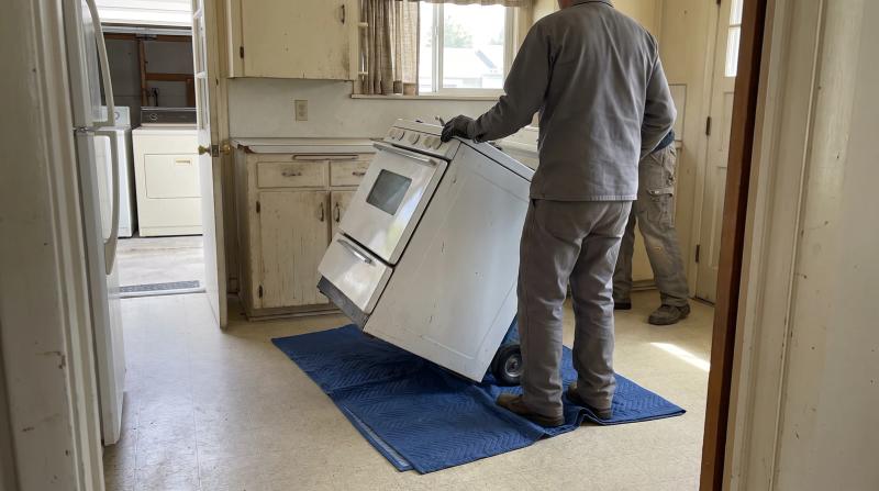 Worker sliding an old white kitchen range onto a moving blanket during a stove removal in a Garden City, Idaho kitchen
