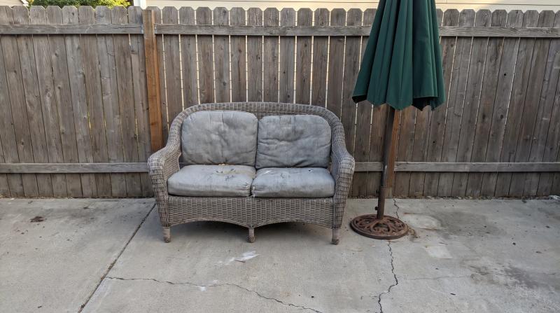 Weathered wicker patio sofa and closed umbrella on a backyard concrete patio in Boise, Idaho, waiting for furniture removal before a home listing