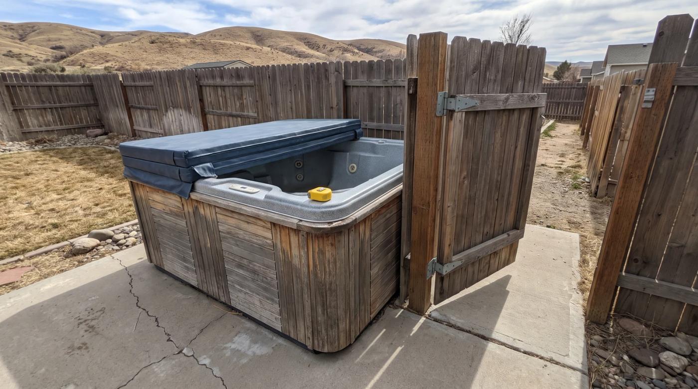 Weathered old hot tub on a backyard patio beside a narrow open side gate in Garden City, Idaho, with a tape measure on the rim