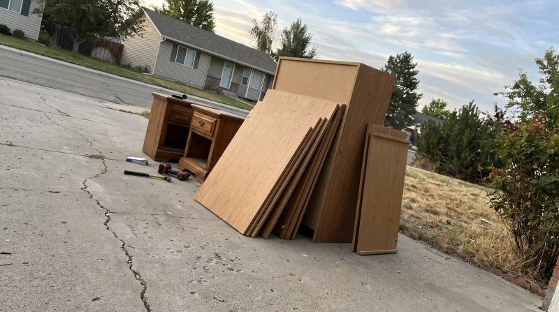 Disassembled laminate dresser panels leaning against two wood nightstands on a driveway during a furniture removal job in Garden City, Idaho.