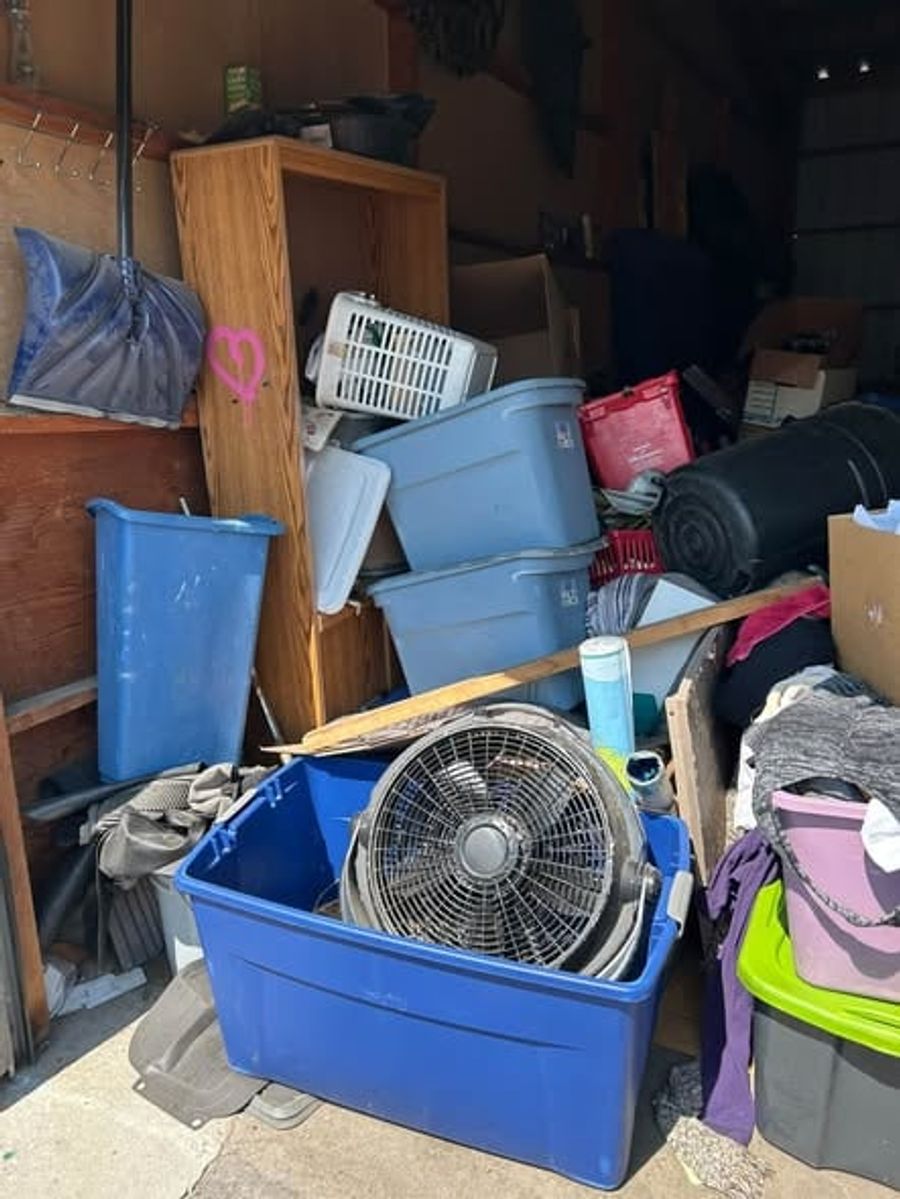 A storage unit packed with plastic totes, a box fan, baskets and bags before a cleanout
