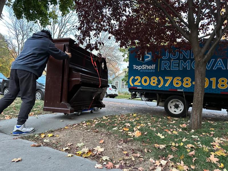 Top Shelf crew pushing an upright piano on a dolly toward the branded truck curbside in Boise Idaho