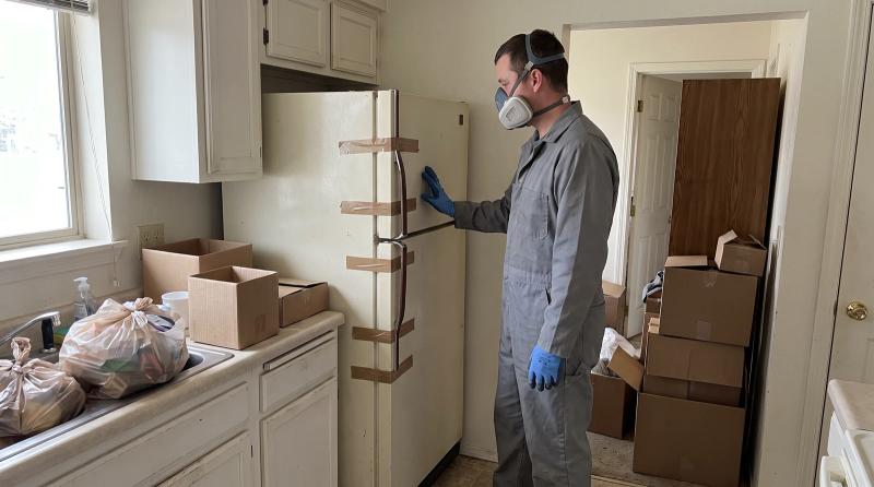 Worker in protective gear next to a taped-shut refrigerator during a hoarding cleanup in a Caldwell, Idaho kitchen