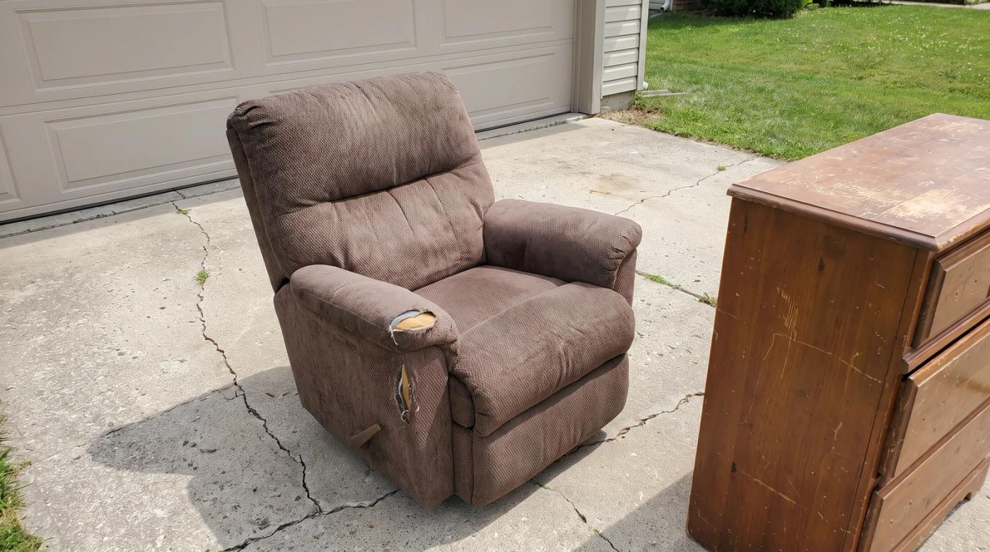 Worn recliner with a torn armrest and a wooden dresser on a driveway in Middleton, Idaho, waiting for donation or furniture removal