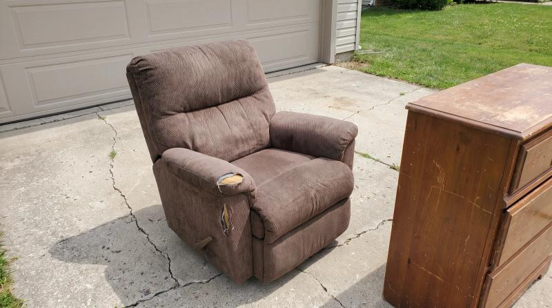 Worn recliner with a torn armrest and a wooden dresser on a driveway in Middleton, Idaho, waiting for donation or furniture removal