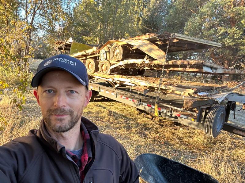 Top Shelf crew member with branded hat standing beside a flatbed trailer full of scrap metal hauled from a rural property in Caldwell Idaho