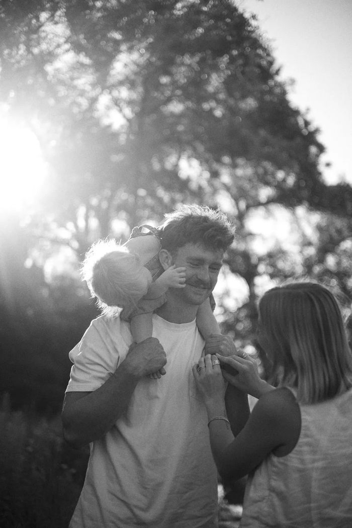 Black-and-white photo of Tommy Simonson with his little one on his shoulders and his wife beside him under the trees