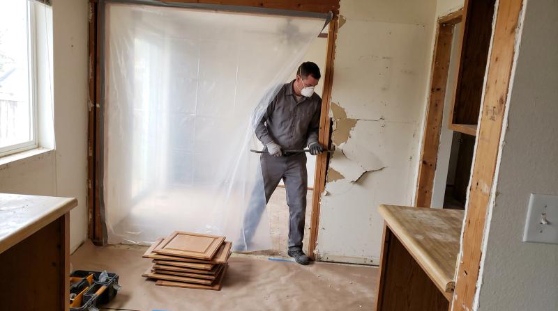 Worker removing kitchen drywall behind a plastic dust barrier during an interior demolition in Meridian, Idaho