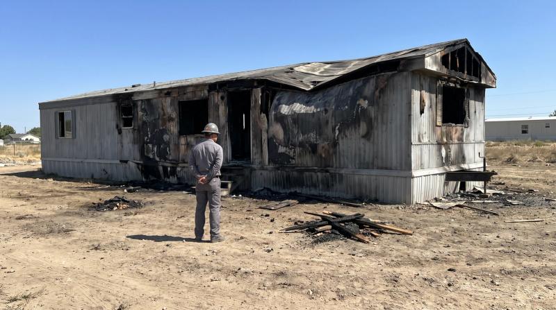 Worker in a hard hat inspecting a fire-damaged single-wide mobile home before demolition on a dirt lot in Meridian, Idaho