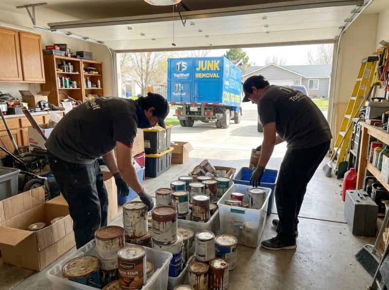 Two Top Shelf crew members sorting and boxing up old paint cans and wood stain in a residential garage with the branded truck visible in Meridian Idaho