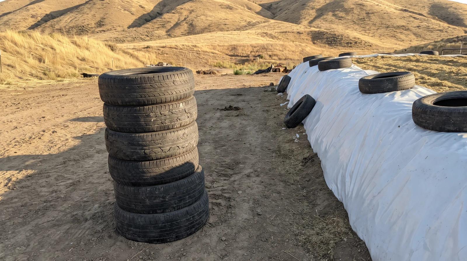Stack of old weathered silage pile tires beside a plastic-covered bunker on a farm near Emmett, Idaho, ready for tire disposal