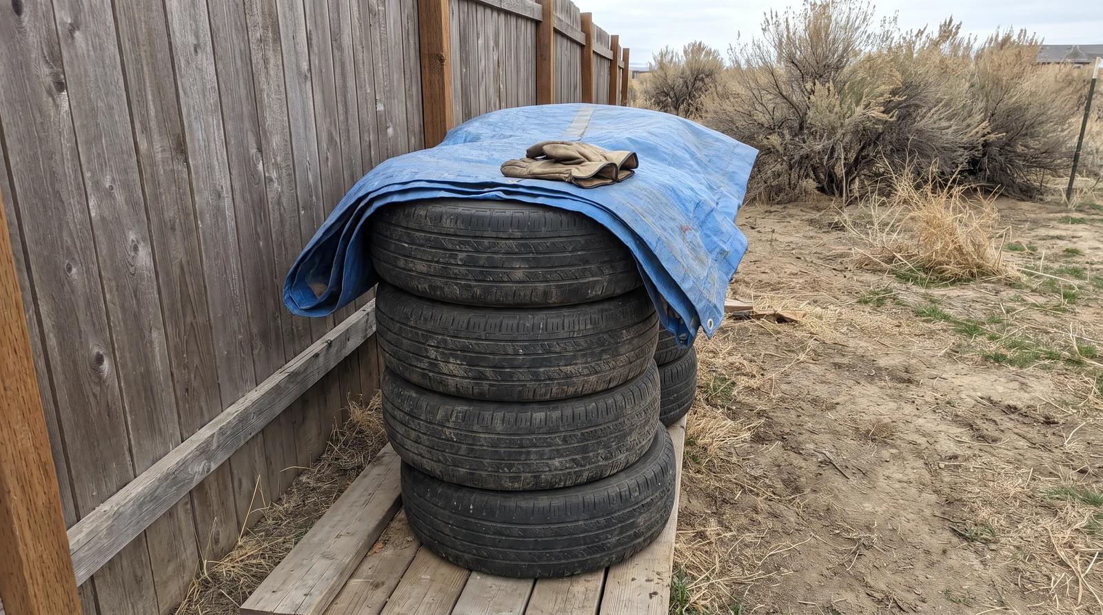 Stack of old tires on boards beside a backyard fence in Garden City, Idaho, partly covered with a blue tarp while they wait for tire disposal
