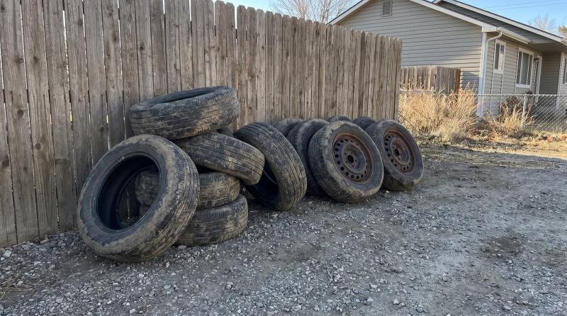 Worn tires stacked on a gravel driveway next to four tires still mounted on steel wheels, with a weathered wooden fence behind them, in Garden City, Idaho