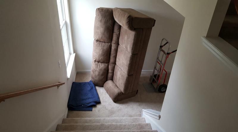 A worn fabric sofa tipped on end at the bottom of a narrow carpeted staircase in an Eagle, Idaho home, with a moving blanket and hand truck nearby
