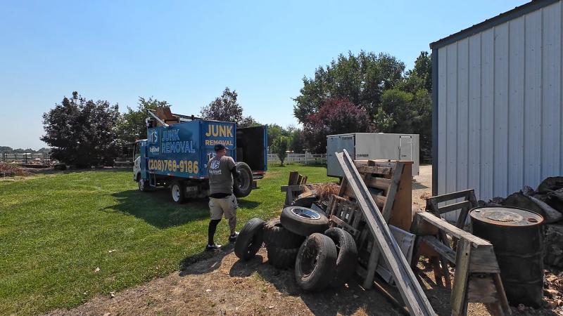 Top Shelf crew member carrying a tire past oil drums and scrap debris to the branded truck at a rural property in Garden City Idaho