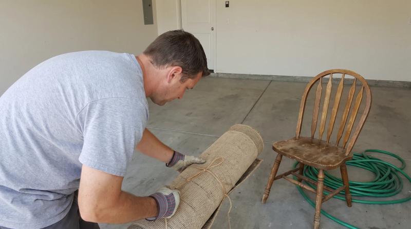 Man in a plain gray T-shirt lifting a rolled-up rug off a swept garage floor during a junk removal job in Meridian, Idaho, with an old wooden chair nearby.