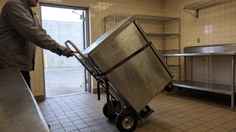 Worker moving an old stainless steel reach-in cooler out of a restaurant kitchen during commercial appliance removal in Nampa, Idaho