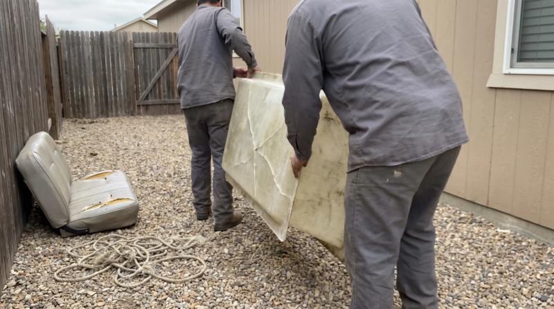 Two workers in plain gray clothes lifting a cracked fiberglass panel from a gravel side yard in Kuna, Idaho, next to a faded vinyl seat cushion and a coil of frayed rope.