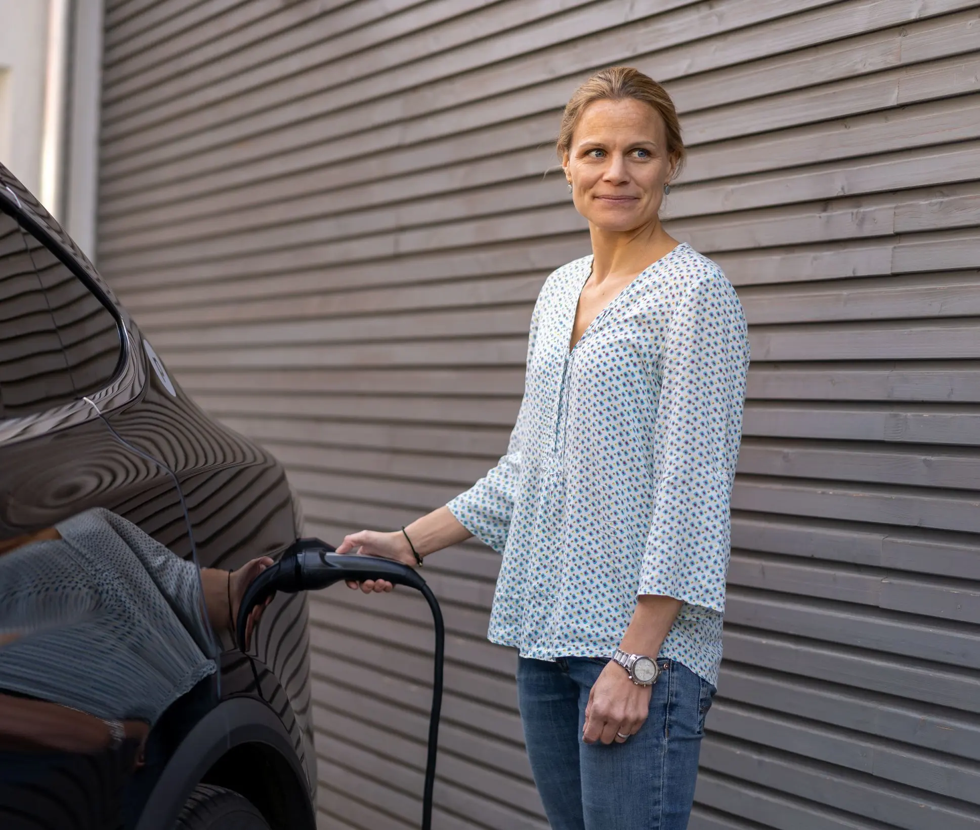 Eine Frau steht neben einer Hauswand und lädt ein E-Auto mit einem Ladekabel