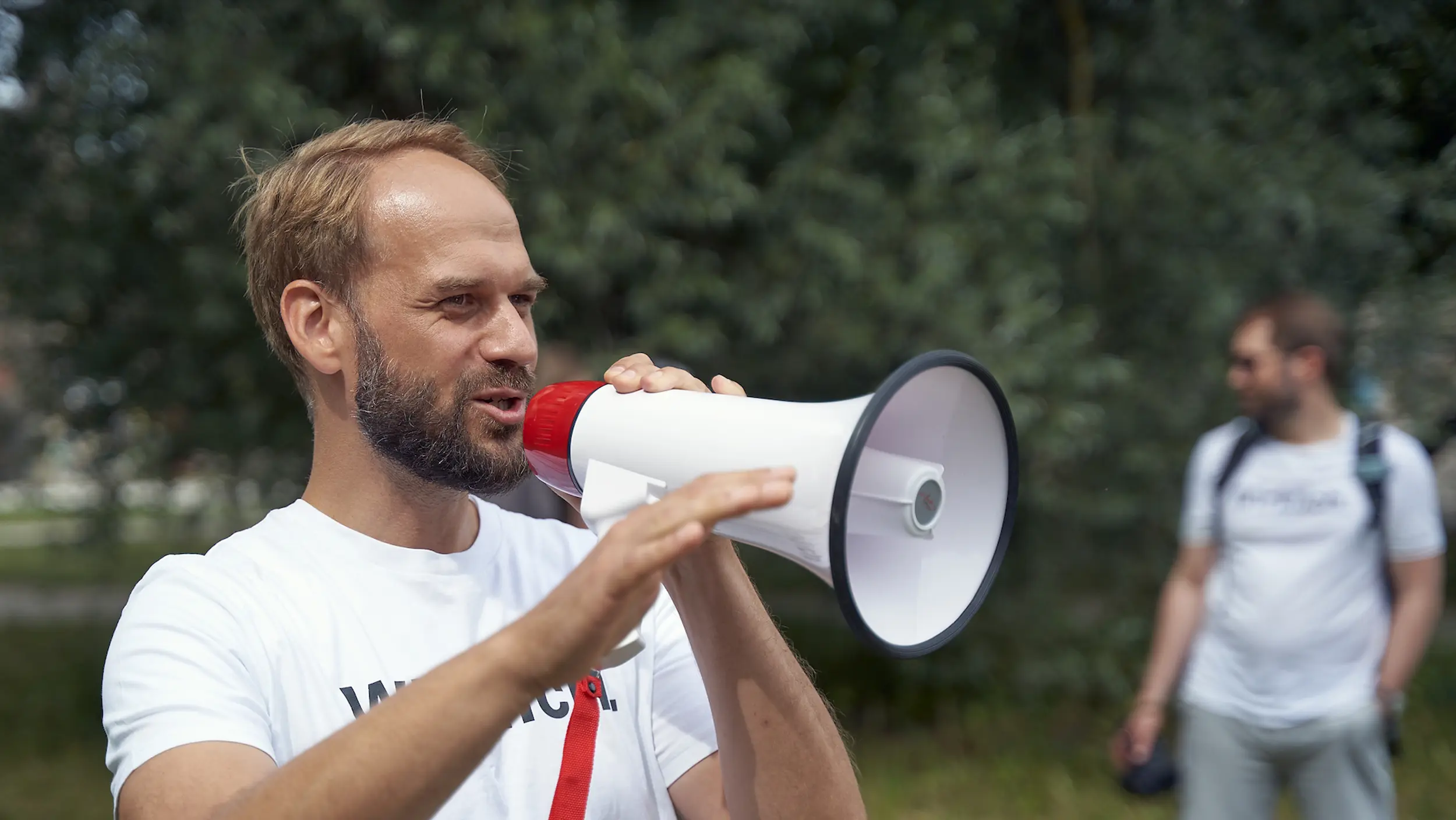 Ein Mann in einem weißen T-Shirt spricht im Freien in ein Megafon.